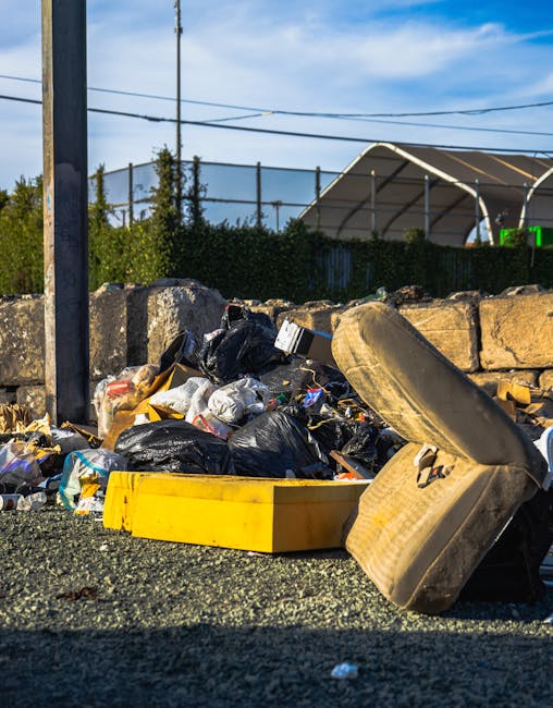 A pile of mixed rubbish and debris is scattered on an asphalt surface near a stone wall. The refuse includes black plastic garbage bags, a yellow plastic container, an old, worn car tyre leaning against the pile, and various loose trash such as paper and packaging. In the background, there is a metal fence topped with greenery, and behind that, a large semi-circular structure with a white roof supports and poles. Overhead, there are horizontal power lines against a blue sky with some clouds. The scene appears to be an informal or unmanaged waste collection area, reflecting private disposal or alternative rubbish removal methods that Rubbish Removal Catford might address, highlighting the importance of proper on-site clearance and waste handling for such urban environments.