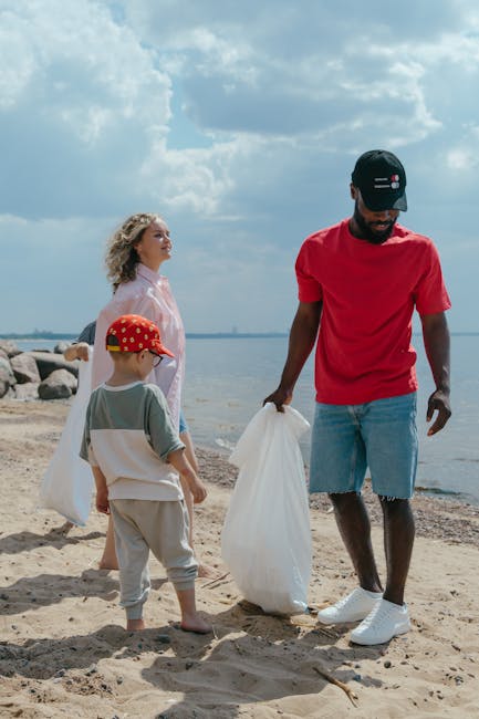 A group of three people, including an adult male and two children, engaged in rubbish removal on a sandy beach near a body of water. The man, wearing a red t-shirt, distressed denim shorts, and a black baseball cap, is holding a white plastic bag while standing on the sand. One child, dressed in a grey t-shirt, beige trousers, and a red cap with yellow patterns, stands nearby holding a similar bag, facing the water. The other child, a girl, with curly blonde hair, wearing a light pink dress, stands slightly behind, observing the scene. In the background, there are large rocks along the shoreline, a calm water surface, and a partly cloudy sky overhead. The scene captures a moment of outdoor waste collection, potentially organized as part of a rubbish removal service, emphasizing on-site cleanup or private disposal efforts in a natural environment, supporting environmental responsibility.