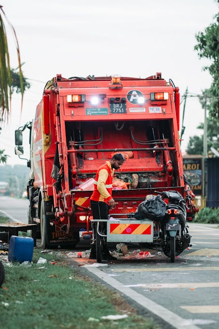 A red metal rubbish skip with visible rust and chipped paint, situated outdoors against a dark green tiled wall on the left and a light grey concrete wall on the right. The skip appears to be partially filled or closed, with a flat lid on top. To the right of the skip, several white plastic rubbish bags, some partially torn open, are piled on the paved ground made of dark interlocking bricks. The bags contain mixed waste, including plastic bottles and paper, indicative of domestic or commercial waste disposal. The setting suggests an urban alleyway or side street area where independent waste collection is taking place, possibly as an alternative to council-led rubbish removal. The lighting is neutral, with no strong shadows, highlighting the textures of the rusted metal, the concrete wall surfaces, and the plastic waste. This scene captures a typical example of private rubbish handling supporting community cleanliness efforts, with the Rubbish Removal Catford service potentially providing removal or disposal solutions for such waste accumulation.