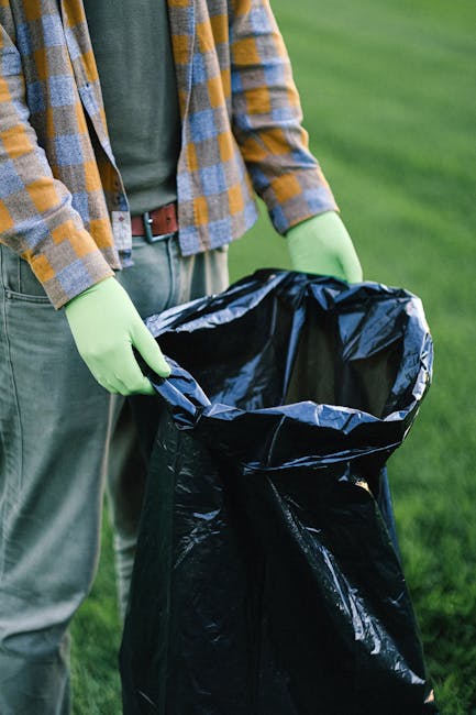 A person wearing a plaid shirt in shades of yellow, blue, and gray, along with gray trousers secured by a brown belt, is seen holding a large black rubbish bag made of coated plastic material. The individual is wearing light green disposable gloves and appears to be in an outdoor environment with a grassy background. The rubbish bag is open at the top, revealing its empty interior, and is positioned upright, being held at the edge to prevent spillage. The scene suggests an activity related to waste collection or disposal, consistent with rubbish removal services. The lighting is natural, indicating daytime, and the overall setting points to an outdoor cleanup or waste management task, typical in garden or outdoor area maintenance, aligned with private or independent collection approaches promoted by services such as Rubbish Removal Catford.
