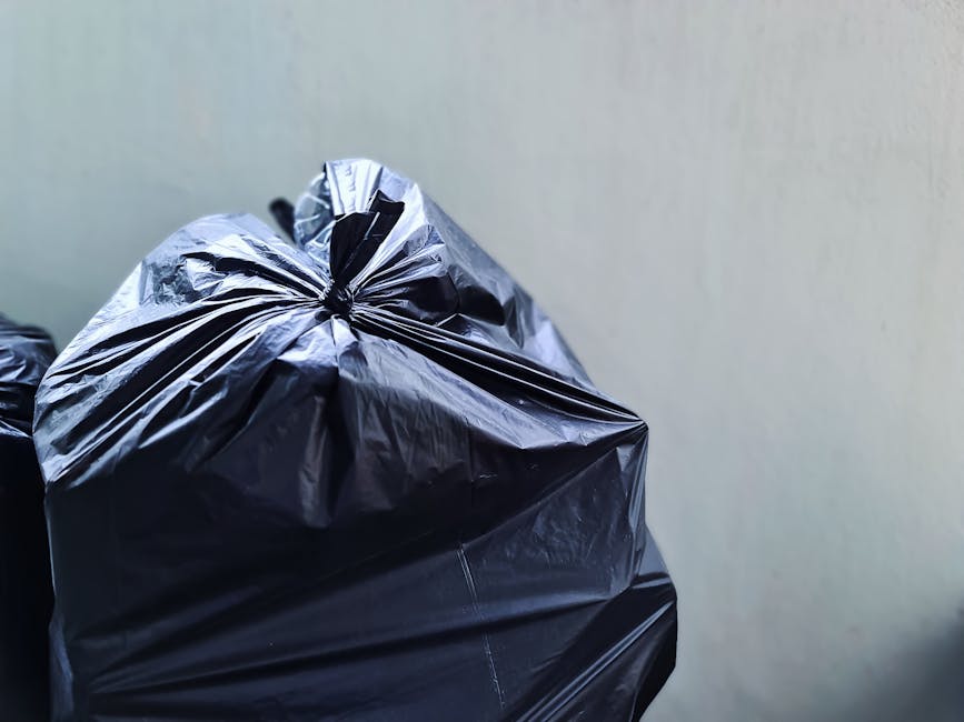 Close-up view of multiple black plastic rubbish bags, tightly knotted at the top, resting against an off-white wall. The bags are made of shiny, smooth plastic material, with some creases and slight wrinkles visible. They appear to be filled with waste, causing their surfaces to be slightly bulging and uneven. The bags are arranged with one prominent bag in the foreground, while others are partially visible behind it. The image suggests an indoor or sheltered setting, possibly an area designated for rubbish collection or disposal. The neutral background and natural lighting highlight the texture and material of the rubbish bags, emphasizing their role in waste removal services. This scene aligns with private waste collection and on-site clearance, typical of services like those offered by Rubbish Removal Catford for efficient rubbish management.