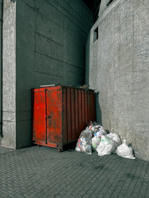 A large pile of black plastic rubbish bags stacked against a modern building with reflective tiles; some bags are torn open, revealing plastic bottles and scattered waste inside. The bags vary in size and are placed directly on a paved surface, with a few leaning or resting atop each other. In the background, graffiti with red spray paint spelling 'XEND' is visible on the tiled wall, and a cloudy sky with patches of orange and gray tones indicates late afternoon or early evening lighting. The scene suggests an area awaiting collection or disposal, consistent with private waste handling or on-site clearance services such as those offered by Rubbish Removal Catford, emphasizing the importance of timely rubbish removal to prevent localised build-up and environmental hazards.