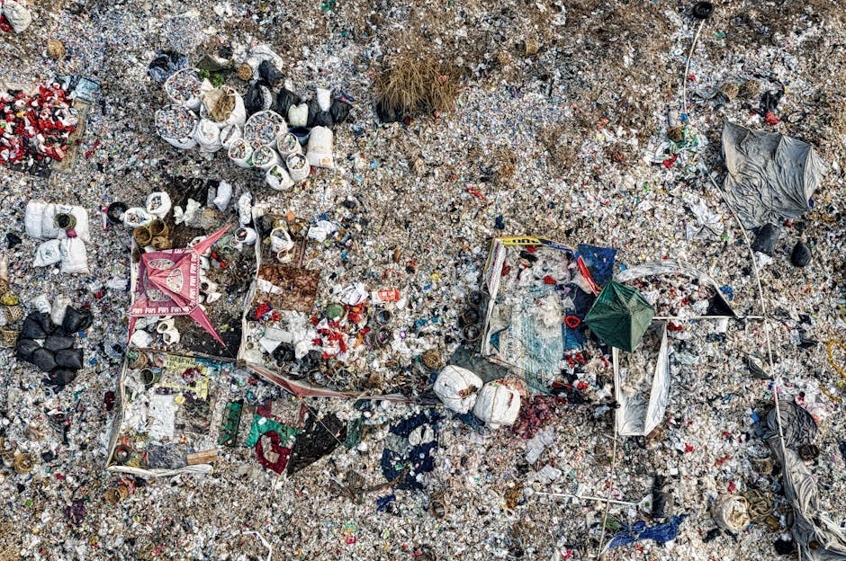 An aerial view of a large, open area filled with various types of waste materials, including crushed cans, plastic bottles, paper, cardboard boxes, and fabric debris, scattered across the ground which appears to be composed of gravel, soil, and small stones. Visible in the scene are piles of garbage—some contained within makeshift barriers made from fabric, plastic sheets, and wooden sticks—while other waste is spread out loosely. The waste features a mix of colours, with metallic silver, white, black, and red hues predominating, alongside some green and transparent plastics. There are no plant or animal elements present, and the environment shows no signs of organized rubbish collection. The scene is illuminated evenly, indicating daylight conditions. The image highlights the extent of unmanaged litter, emphasizing the importance of proper waste disposal services such as those offered by Rubbish Removal Catford for private or independent waste clearance solutions around Rushey Green Catford.